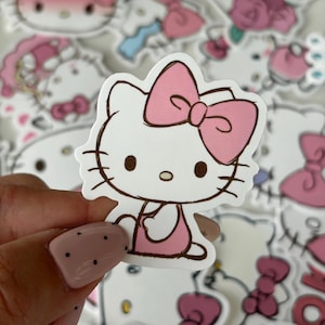 Può includere: Un adesivo fustellato raffigurante un personaggio gatto bianco dei cartoni animati con un grande fiocco rosa sulla testa, occhi neri, un piccolo naso giallo e contorni marroni. Il personaggio indossa un vestito rosa su uno sfondo di adesivi simili.