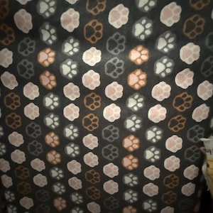 May include: Dark fabric featuring a repeating pattern of paw prints in pink, brown, white, and grey. The fabric has the text "Not Intended for Children's Sleepwear" printed on it.
