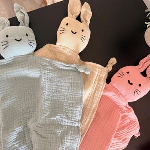 May include: Three soft, bunny-shaped baby security blankets in pastel colors: light blue, beige, and pink. Each bunny head has embroidered facial features and long ears. The blankets are made of a textured, crinkled fabric.