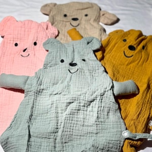 May include: A collection of five soft, bear-shaped baby blankets in various colors: pink, tan, blue-green, and yellow. Each blanket features embroidered facial details and attached arms. The blankets are made of a textured fabric.