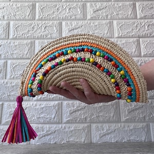 May include: A woven straw clutch with a rainbow beaded design and a colorful tassel. The clutch features a half-circle shape with orange, blue, and yellow accents. The tassel is made of pink, orange, and teal strands.