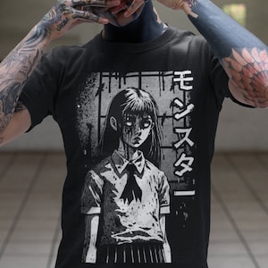 May include: A black t-shirt with a graphic print of a girl with long black hair and a white collar. The girl is in a white and black school uniform. The text "モジズメ" is printed on the right side of the shirt.