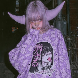May include: A long-sleeved lavender sweatshirt featuring an all-over print of small teddy bears and adhesive bandages. The center graphic displays a stylized illustration of a person with dark hair, a spiked collar, and Japanese text.