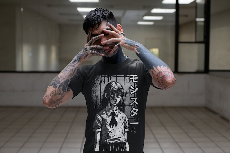 May include: A black t-shirt with a graphic print of a girl with long black hair and a white collar. The girl is in a white and black school uniform. The text "モジズメ" is printed on the right side of the shirt.