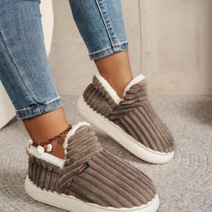 May include: Brown corduroy slippers with white faux fur trim and white soles. The slippers have a high-top design and a sporty look. The corduroy has vertical ridges. The slippers are worn with blue jeans.