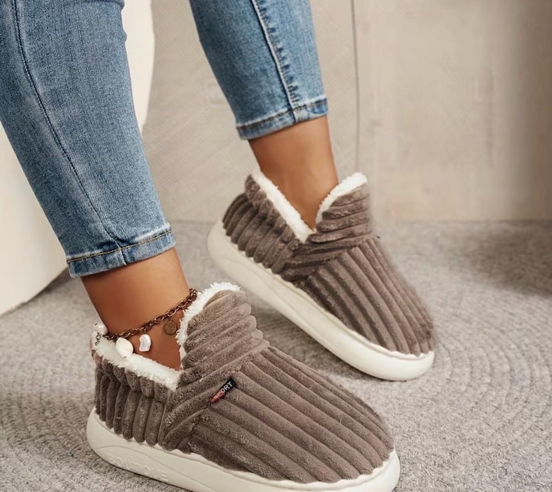 May include: Brown corduroy slippers with white faux fur trim and white soles. The slippers have a high-top design and a sporty look. The corduroy has vertical ridges. The slippers are worn with blue jeans.