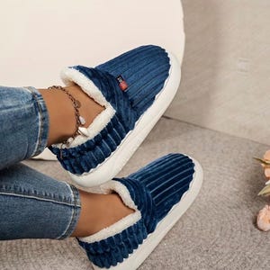 May include: A pair of blue corduroy slippers with white faux fur lining and soles. The slippers have a slip-on design and a small red tag that says "Sport". The slippers are designed for indoor wear.