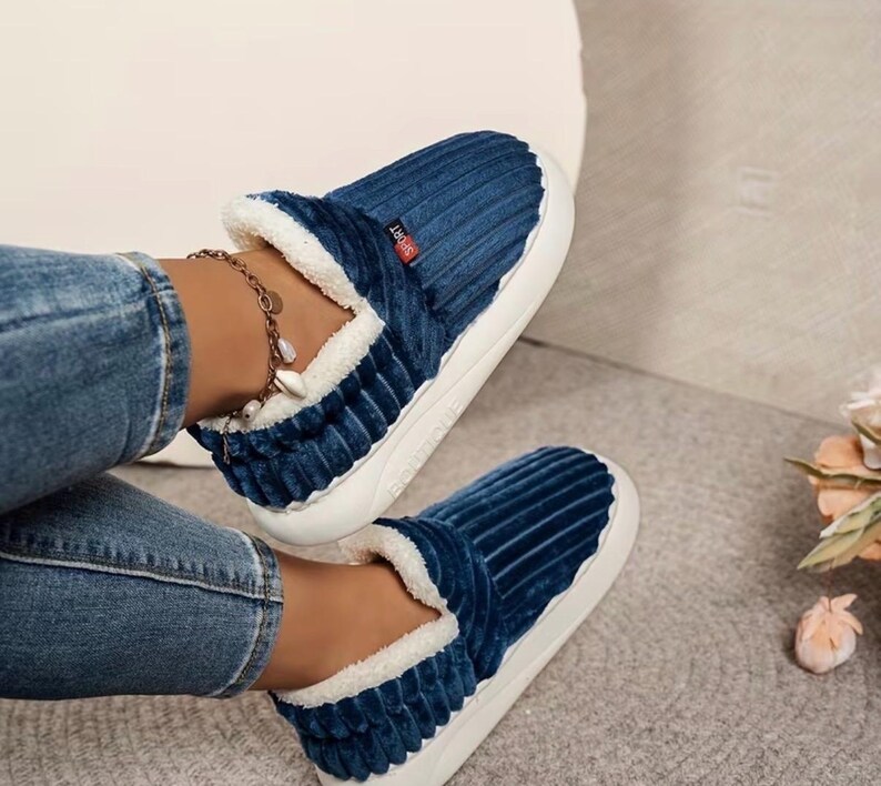 May include: A pair of blue corduroy slippers with white faux fur lining and soles. The slippers have a slip-on design and a small red tag that says "Sport". The slippers are designed for indoor wear.