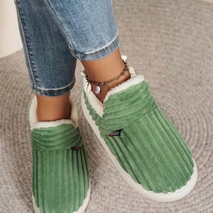 May include: Green corduroy slippers with a white, fluffy interior and a white sole. The slippers have a small "SPORT" tag on the side. The image shows the slippers being worn with blue jeans.