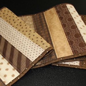May include: A quilted table runner in shades of brown and beige. The runner features a patchwork design with various patterns, including stripes, polka dots, and floral motifs. The edges are neatly finished with a brown border, creating a decorative accent for any table.