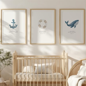 May include: Three framed prints with nautical themes hang above a wooden crib. The prints feature an anchor, a life preserver, and a whale, each with a corresponding inspirational quote. The room also includes a wicker chair and a woven basket.