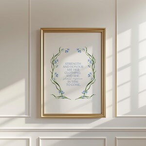 May include: A framed print with a gold-colored frame. The artwork features a floral wreath in blue and green, surrounding text that reads "STRENGTH AND HONOUR ARE HER CLOTHING; AND SHE shall rejoice IN TIME TO COME."