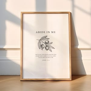 Op de afbeelding: Een zwart-witte illustratie van een olijftak met de tekst "ABIDE IN ME" en de Bijbeltekst "Johannes 15:4" onder de afbeelding.