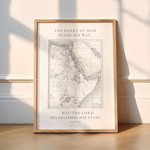 May include: A framed print featuring a monochrome vintage map. The text at the top says "THE HEART OF MAN PLANS HIS WAY," and the bottom says "BUT THE LORD ESTABLISHES HIS STEPS." The frame is made of light-coloured wood.
