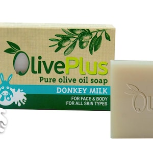 Könnte beinhalten: Ein Stück OlivePlus Eselsmilchseife in hellem Beige. Daneben eine Schachtel mit dem OlivePlus-Logo, einer Olivenzweig-Illustration und dem Text: "Pure olive oil soap, Donkey Milk, For Face & Body, For All Skin Types."
