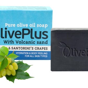 Könnte beinhalten: Ein dunkelgraues Seifenstück und eine Seifenschachtel mit dem Text "Pure olive oil soap LivePlus With Volcanic sand & SANTORINI'S GRAPES". Eine Traube grüner Trauben und Blätter befinden sich vor der Schachtel. Die Seife ist für alle Hauttypen.