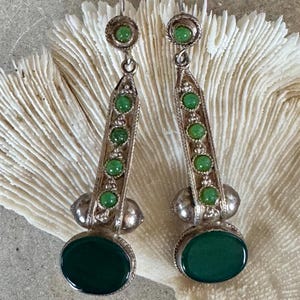 Pu&ograve; includere: Un paio di orecchini color argento con un design pendente. Ogni orecchino presenta una grande pietra ovale verde scuro nella parte inferiore, con pietre verdi pi&ugrave; piccole e dettagli decorativi argentati. Chiusura a gancio.