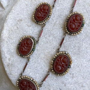 May include: A vintage necklace featuring oval, reddish-brown cameos with floral carvings. Each cameo is framed with small, off-white beads and connected by silver-toned links. The necklace is displayed on a light-colored surface.