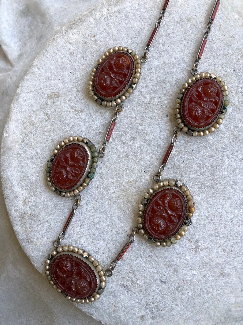 May include: A vintage necklace featuring oval, reddish-brown cameos with floral carvings. Each cameo is framed with small, off-white beads and connected by silver-toned links. The necklace is displayed on a light-colored surface.