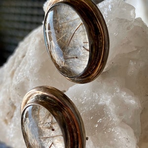 Puede incluir: Un par de pendientes de bot&oacute;n con una parte superior de cristal transparente y abovedada, que muestra delicadas inclusiones en forma de cabello. Los pendientes tienen un engaste de metal plateado con una banda oscura y texturizada. Se exhiben sobre una superficie cristalina blanca.