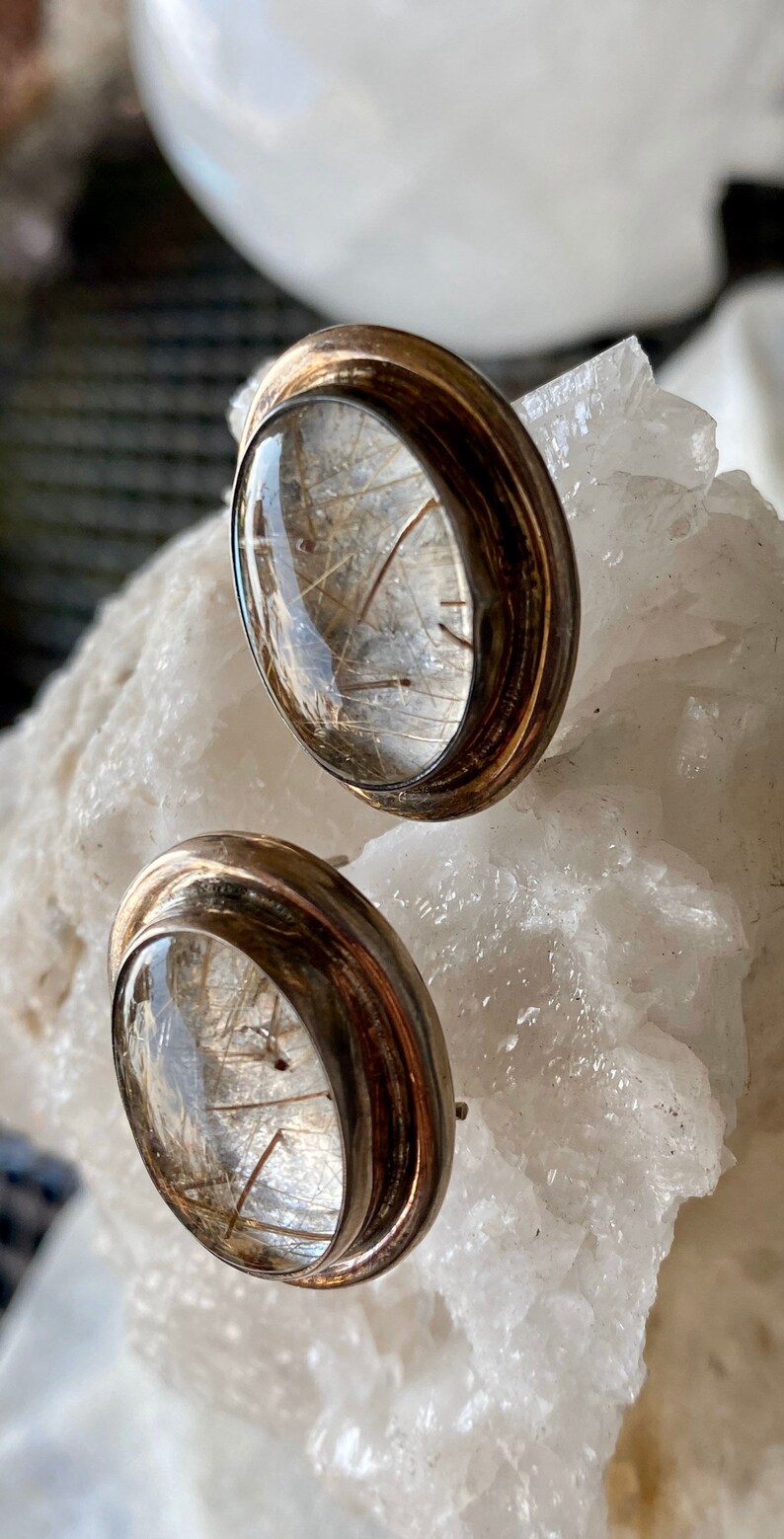 Puede incluir: Un par de pendientes de bot&oacute;n con una parte superior de cristal transparente y abovedada, que muestra delicadas inclusiones en forma de cabello. Los pendientes tienen un engaste de metal plateado con una banda oscura y texturizada. Se exhiben sobre una superficie cristalina blanca.