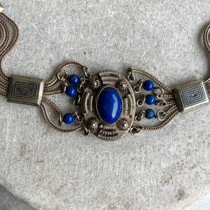 May include: A silver chain bracelet with a large blue lapis lazuli stone set in a silver filigree design. The bracelet has multiple strands of chain and small blue beads.