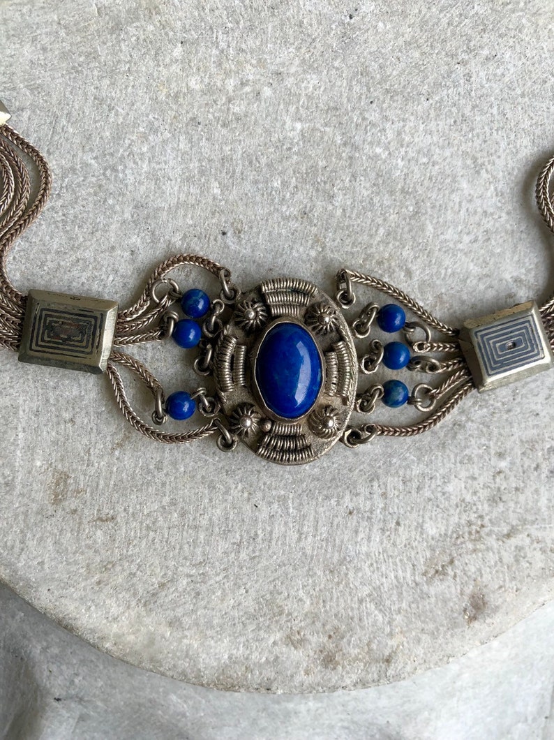 May include: A silver chain bracelet with a large blue lapis lazuli stone set in a silver filigree design. The bracelet has multiple strands of chain and small blue beads.