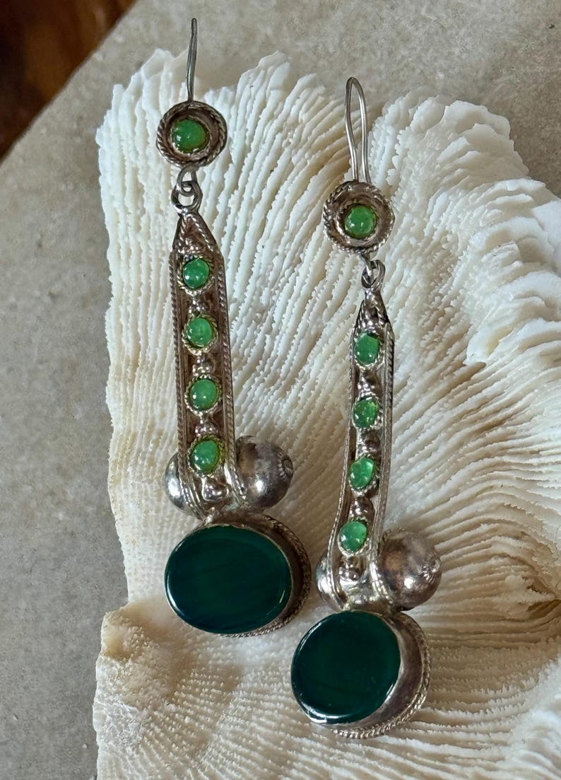 Pu&ograve; includere: Orecchini color argento con pietre verdi. Ogni orecchino presenta una pietra verde circolare in alto, una serie di pietre verdi pi&ugrave; piccole al centro e una grande pietra verde ovale in basso. Design vintage.