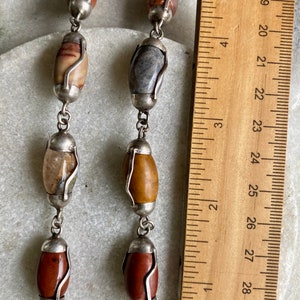 May include: A silver chain necklace with oval-shaped gemstone beads in various colors, including brown, gray, and white. The necklace is measured against a ruler, showing it is approximately 26 inches long.