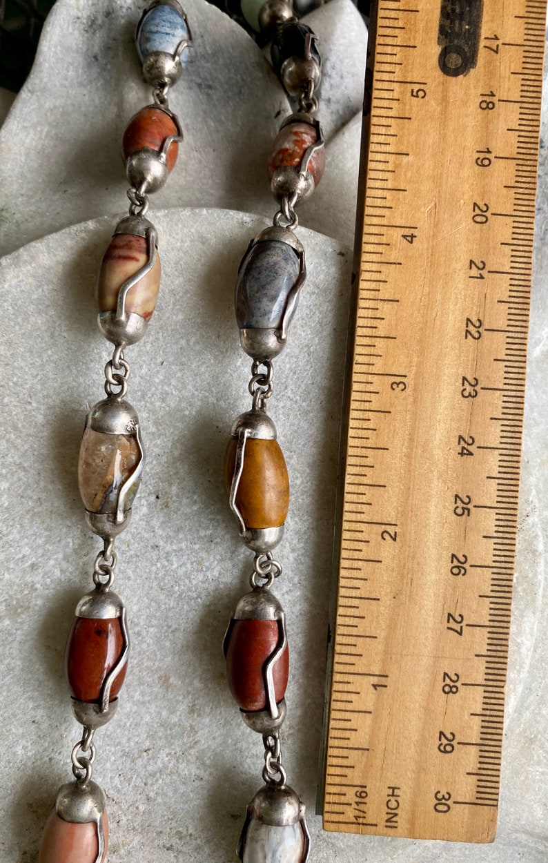 May include: A silver chain necklace with oval-shaped gemstone beads in various colors, including brown, gray, and white. The necklace is measured against a ruler, showing it is approximately 26 inches long.