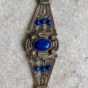 May include: A silver bracelet with a large blue lapis lazuli stone set in a decorative silver frame. The bracelet has multiple strands of silver chain and blue lapis lazuli beads.