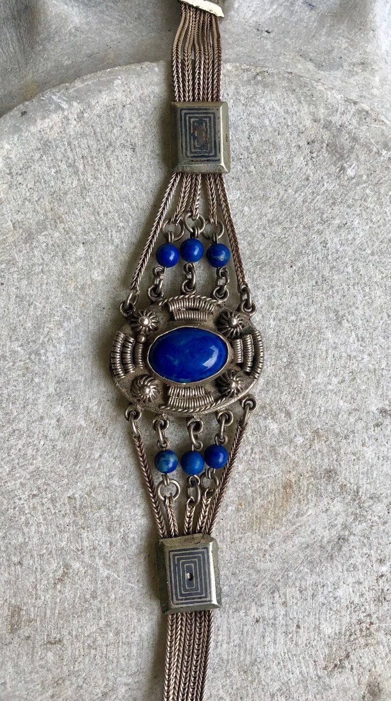 May include: A silver bracelet with a large blue lapis lazuli stone set in a decorative silver frame. The bracelet has multiple strands of silver chain and blue lapis lazuli beads.