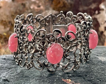 Puede incluir: Un brazalete de filigrana plateado con cuatro piedras ovaladas de color rosa. El brazalete está abierto y tiene un cierre de bisagra.
