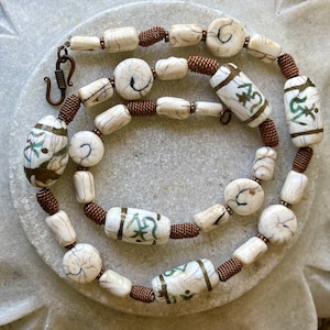 May include: A white and brown beaded necklace with copper wire accents. The beads are decorated with gold and green symbols.