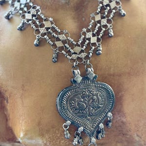 May include: A silver-toned necklace with a large heart-shaped pendant. The pendant features an embossed design with two birds. The necklace has multiple strands with diamond-shaped links and small dangling charms. The necklace is displayed on a neutral background.