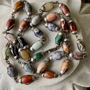 May include: A multi-colored gemstone necklace with silver accents. The necklace features oval-shaped stones in various colors, including brown, red, green, blue, and white. The stones are set in silver frames and connected by a silver chain.