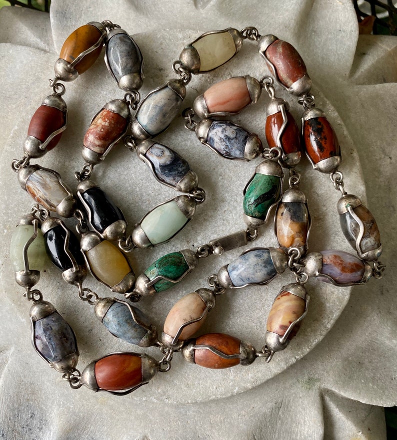 May include: A multi-colored gemstone necklace with silver accents. The necklace features oval-shaped stones in various colors, including brown, red, green, blue, and white. The stones are set in silver frames and connected by a silver chain.
