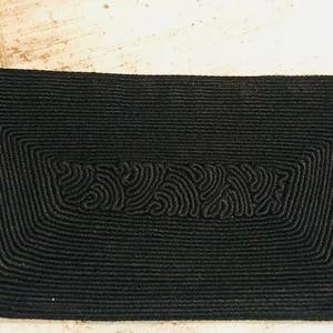 May include: A black woven clutch bag with a gold zipper pull. The bag has a textured design with a repeating pattern.