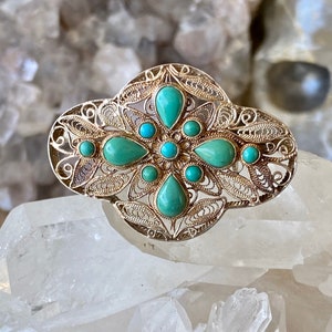 May include: A silver filigree brooch featuring a turquoise stone design. The brooch is shaped like a flower with a central turquoise stone and smaller turquoise stones surrounding it.