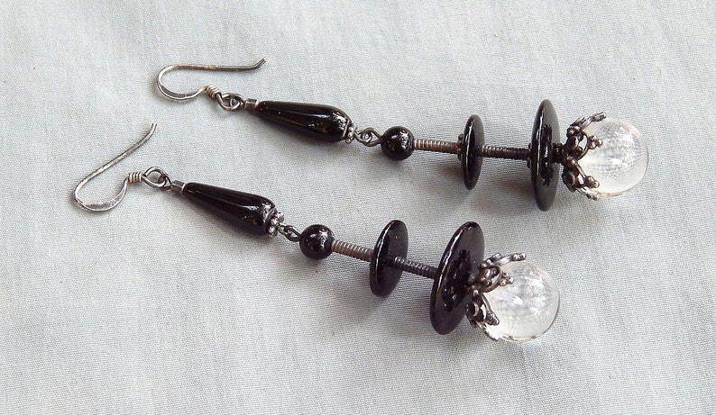 May include: A pair of silver earrings with black and clear glass beads. The earrings feature a teardrop-shaped bead, a round bead, and a clear glass orb.