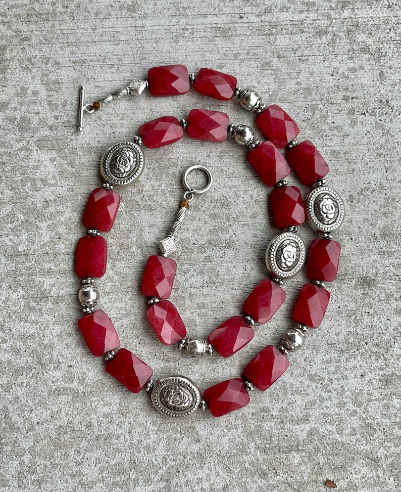 May include: A red gemstone necklace with silver accents. The necklace features faceted, rectangular red gemstones and silver oval charms with a floral design.