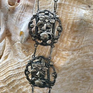 May include: A silver chain with decorative links featuring floral designs. Each link is intricately crafted with a raised floral motif and a small, round bead at each corner.