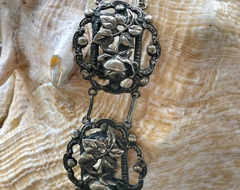 May include: A silver chain with decorative links featuring floral designs. Each link is intricately crafted with a raised floral motif and a small, round bead at each corner.