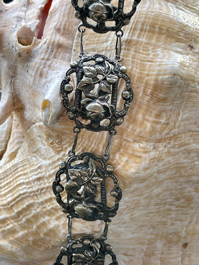 May include: A silver chain with decorative links featuring floral designs. Each link is intricately crafted with a raised floral motif and a small, round bead at each corner.