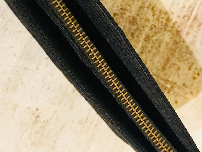 May include: Close-up of a black zipper with gold teeth. The zipper is sewn into a black fabric.