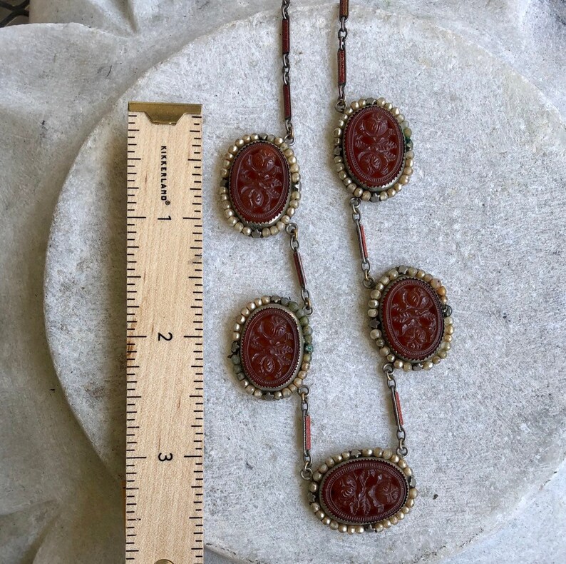 May include: A vintage necklace with five oval, reddish-brown cameo pendants. Each cameo features a floral design and is framed by small, off-white beads. The necklace is approximately 3 inches long, as measured by a ruler.