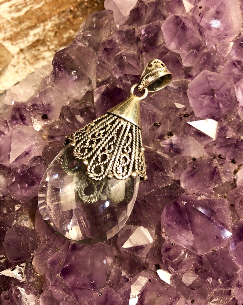 以下が含まれることがあります： 銀のフィリグリーデザインが施された透明な水晶のペンダント。アメジストの結晶のベッドの上に置かれています。