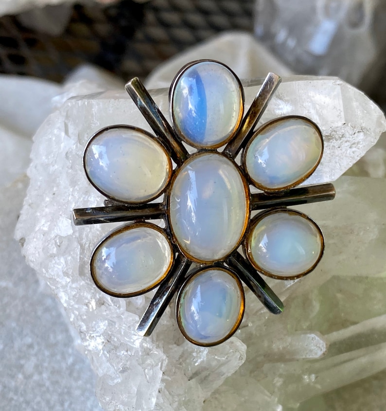 K&ouml;nnte beinhalten: Eine silberne Brosche in Blumenform mit sieben schillernden wei&szlig;en Mondstein-Cabochons, die in einem Sternburst-Design gefasst sind.