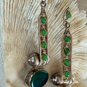 Pu&ograve; includere: Orecchini color argento con pietre preziose verdi. Ogni orecchino presenta un design lungo e rettangolare con una fila di piccole pietre verdi e una pietra verde ovale pi&ugrave; grande nella parte inferiore. Una piccola pietra verde sovrasta ogni orecchino.