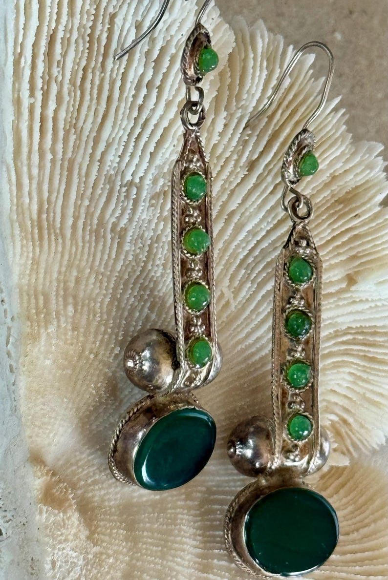 Pu&ograve; includere: Orecchini color argento con pietre preziose verdi. Ogni orecchino presenta un design lungo e rettangolare con una fila di piccole pietre verdi e una pietra verde ovale pi&ugrave; grande nella parte inferiore. Una piccola pietra verde sovrasta ogni orecchino.